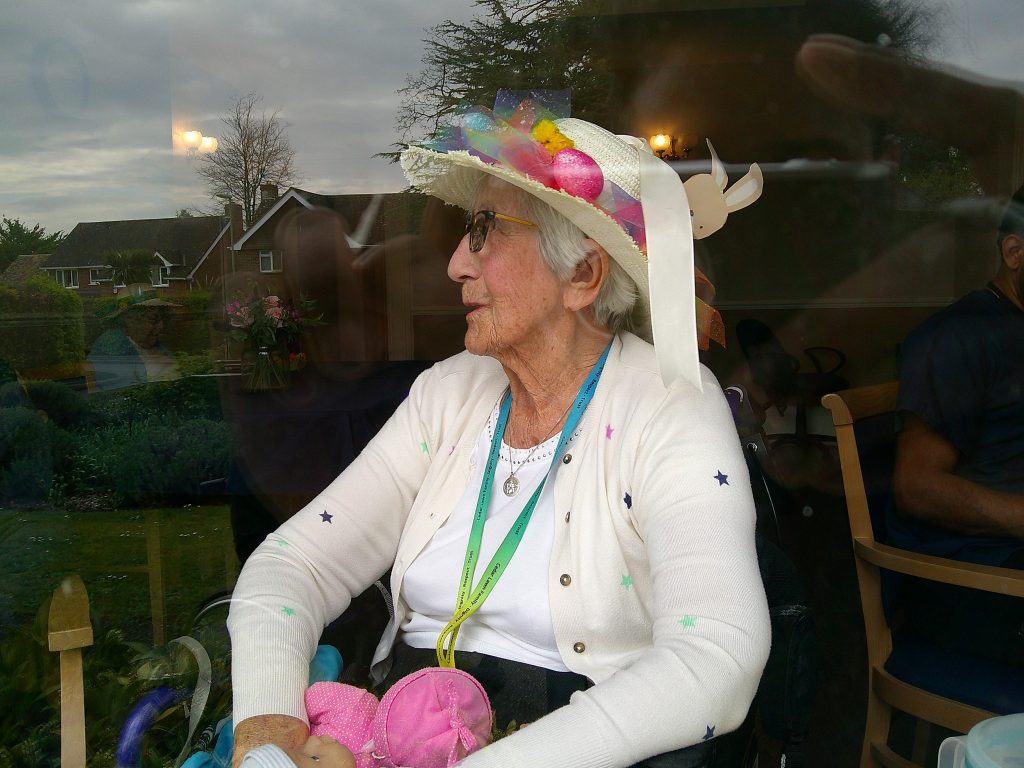 Elderly woman wearing a colorful decorated hat, sitting indoors and looking out through a window.