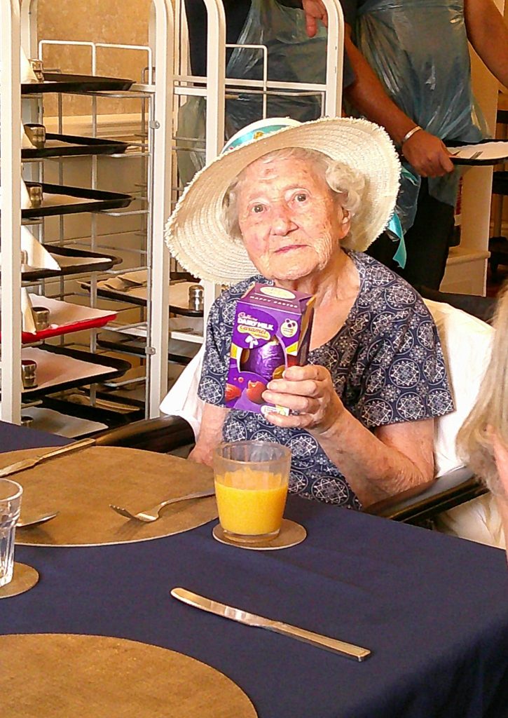 Elderly woman wearing a sun hat sitting at a table, holding a small drink carton.