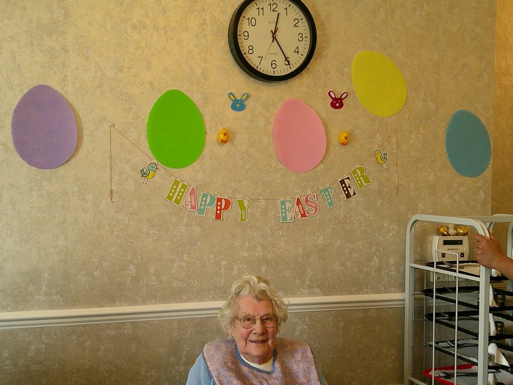 Elderly woman sitting beneath colorful “Happy Easter” decorations on a wall.