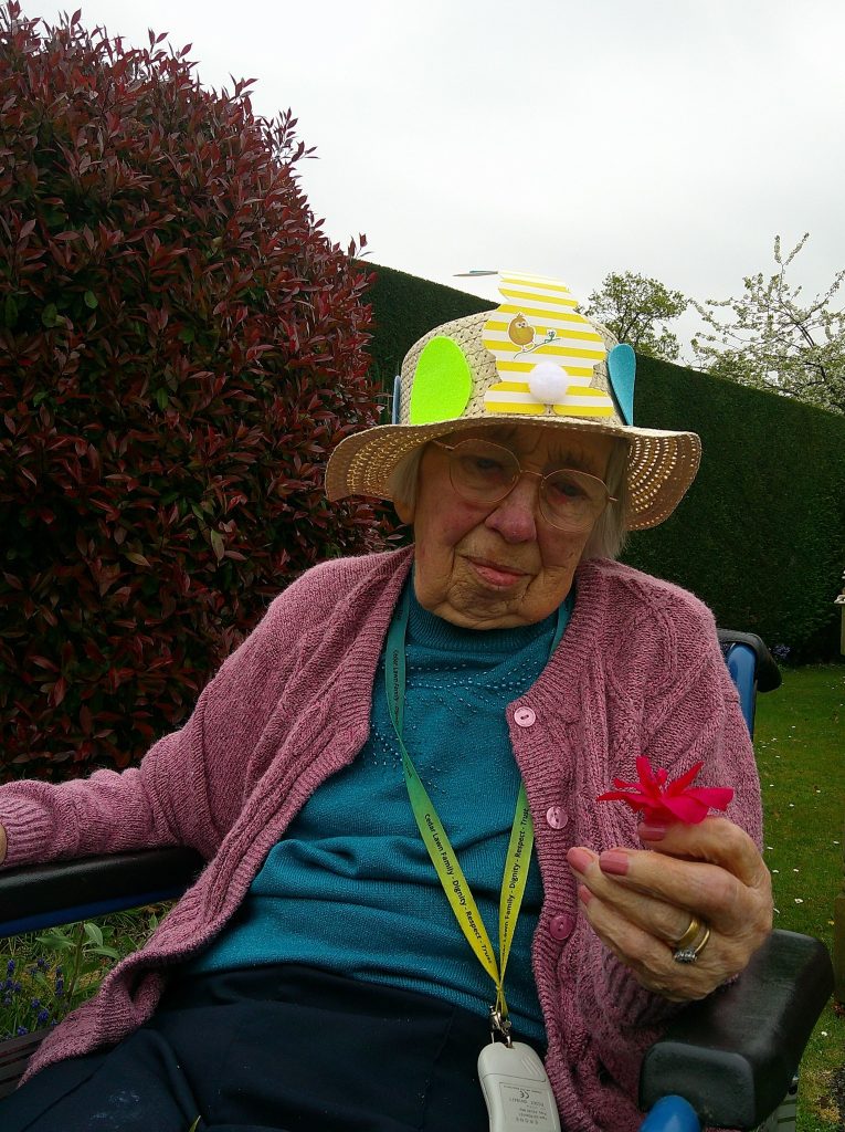 Elderly woman wearing a decorated Easter hat, sitting outdoors and holding a bright pink flower.