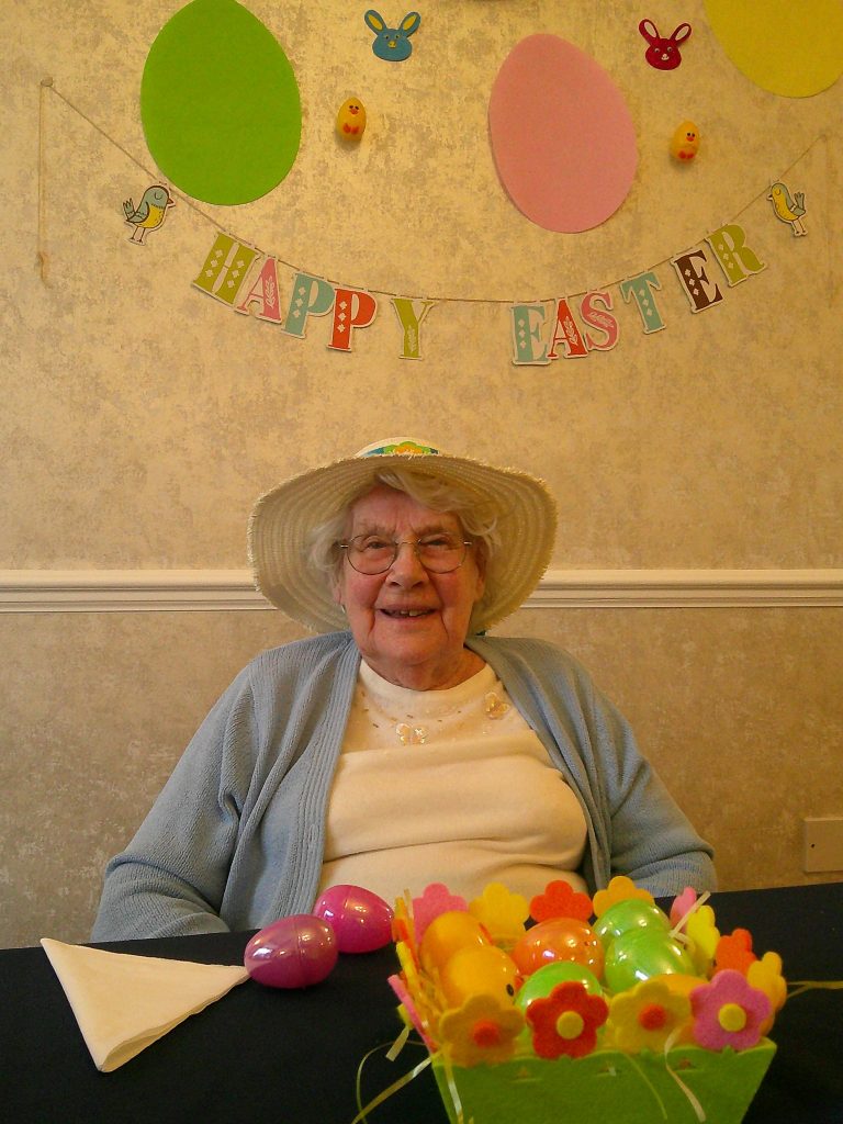 Elderly woman wearing a hat, sitting at a table with colorful Easter eggs and decorations under a “Happy Easter” banner.
