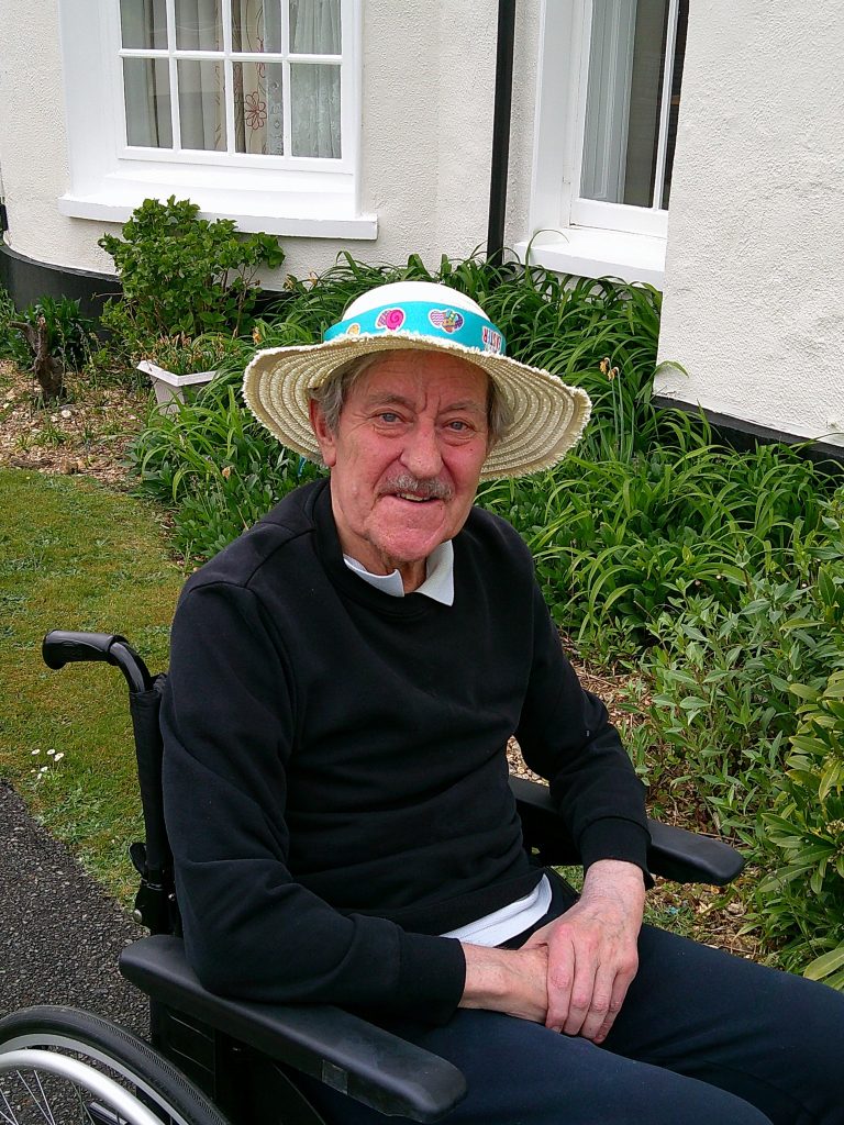 Elderly man in a wheelchair wearing a decorated straw hat, smiling outdoors in a garden near a building.