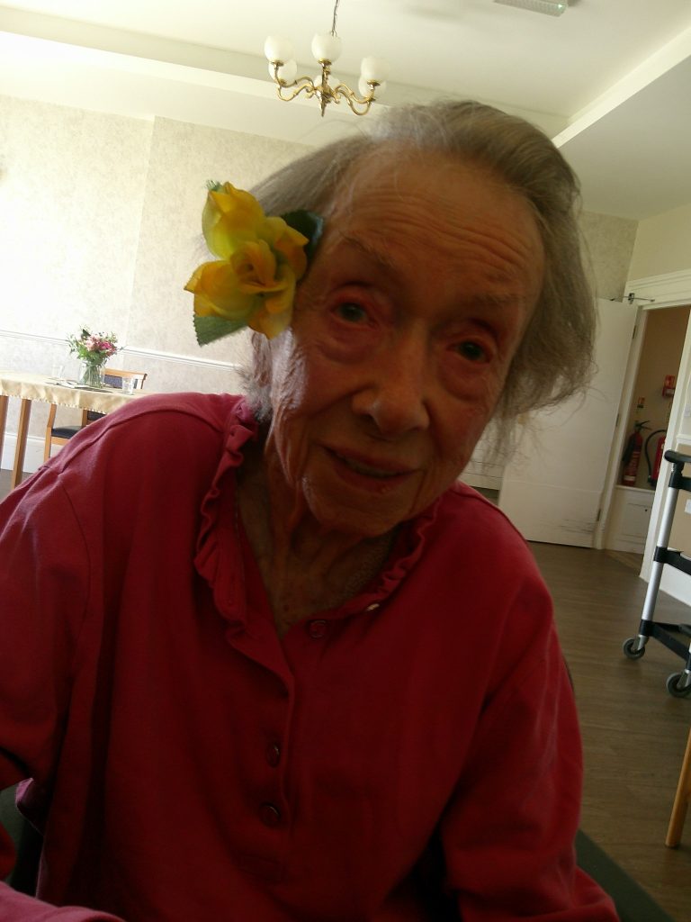 Elderly woman in a red top smiling indoors with a yellow flower in her hair.