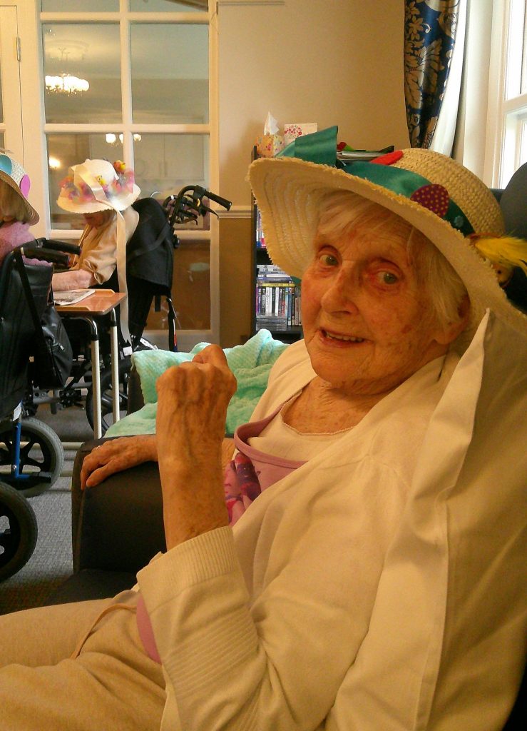 Elderly woman wearing a decorated hat sits in a chair, smiling indoors, with other seniors in the background.