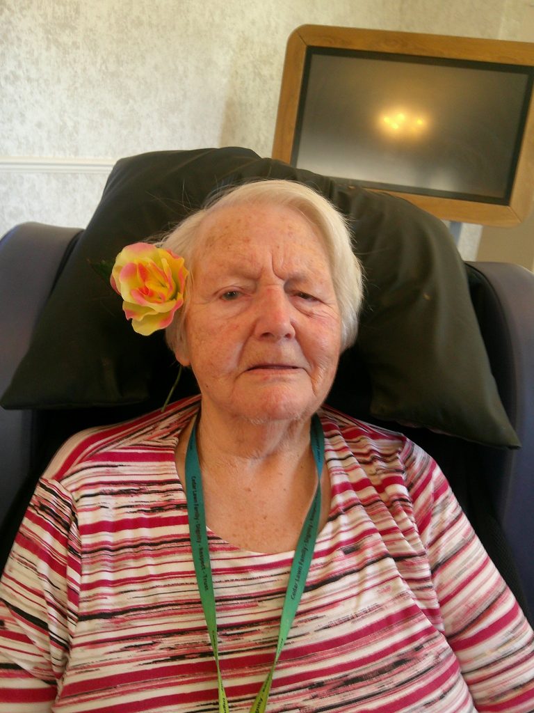 Elderly woman sitting in a chair indoors, wearing a striped top and a flower tucked behind her ear.