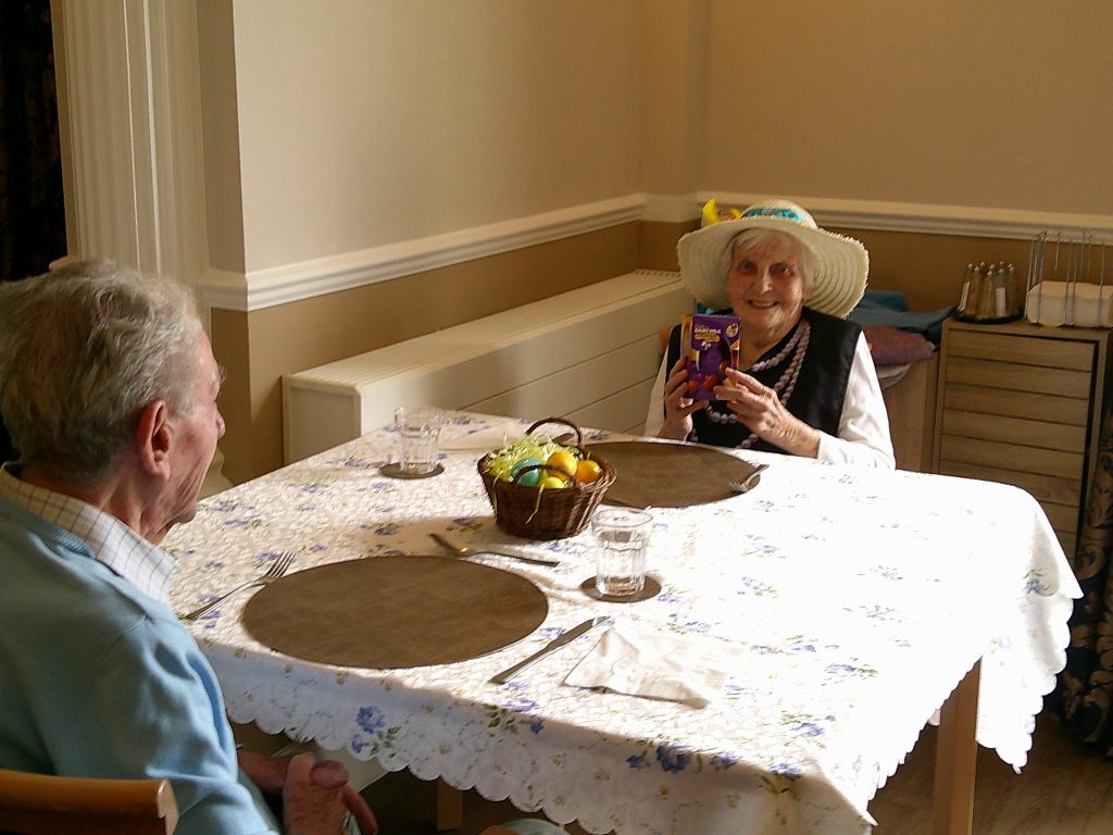Two elderly people sitting at a table, with one holding a small gift box and smiling.