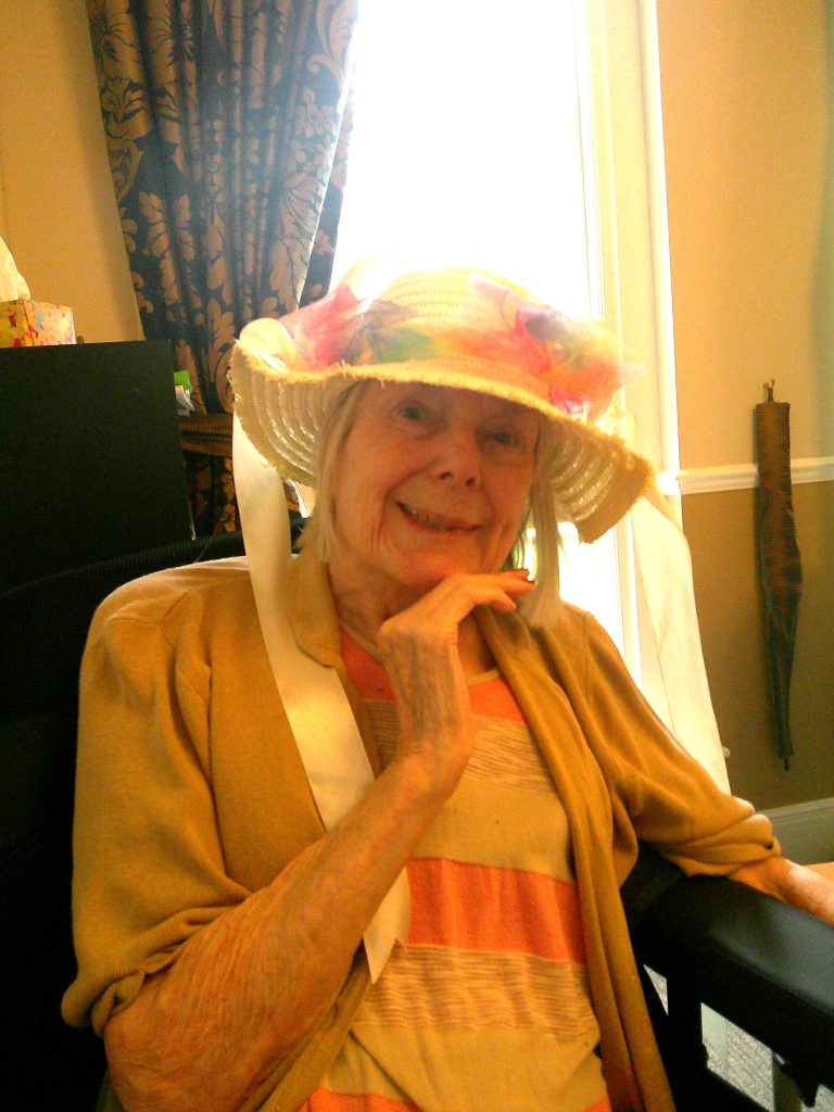 Elderly woman smiling indoors, wearing a colorful decorated hat and sitting by a bright window.