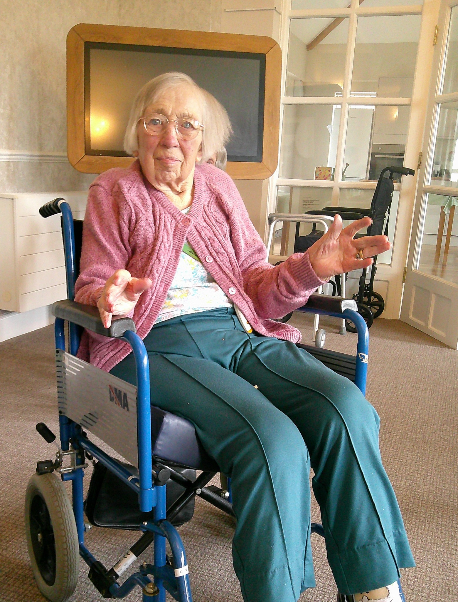 Elderly woman in a wheelchair gestures with her hands while sitting indoors in a care facility.