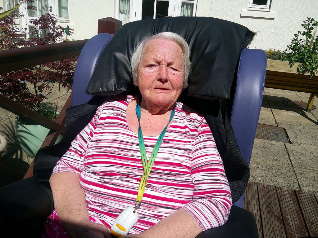Elderly woman sitting in a cushioned chair outdoors in the sun, wearing a striped top and lanyard.