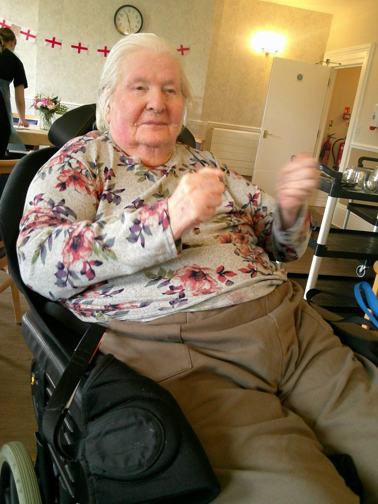 Elderly woman sitting in a chair indoors, moving her hands, with festive decorations in the background.