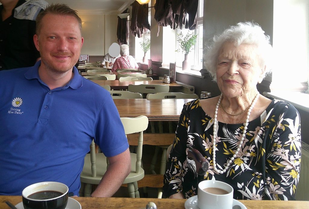An elderly woman and a man sit together at a café table, smiling with cups of coffee in front of them.