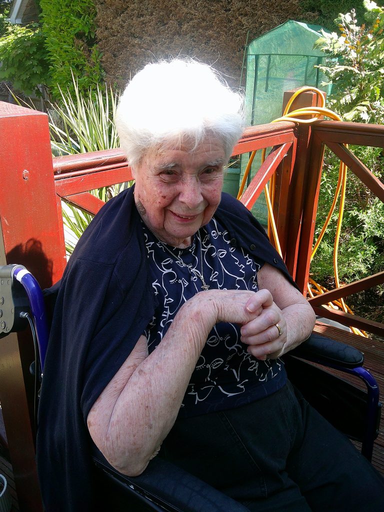 Smiling elderly woman sitting outdoors in a wheelchair, enjoying a sunny garden setting.