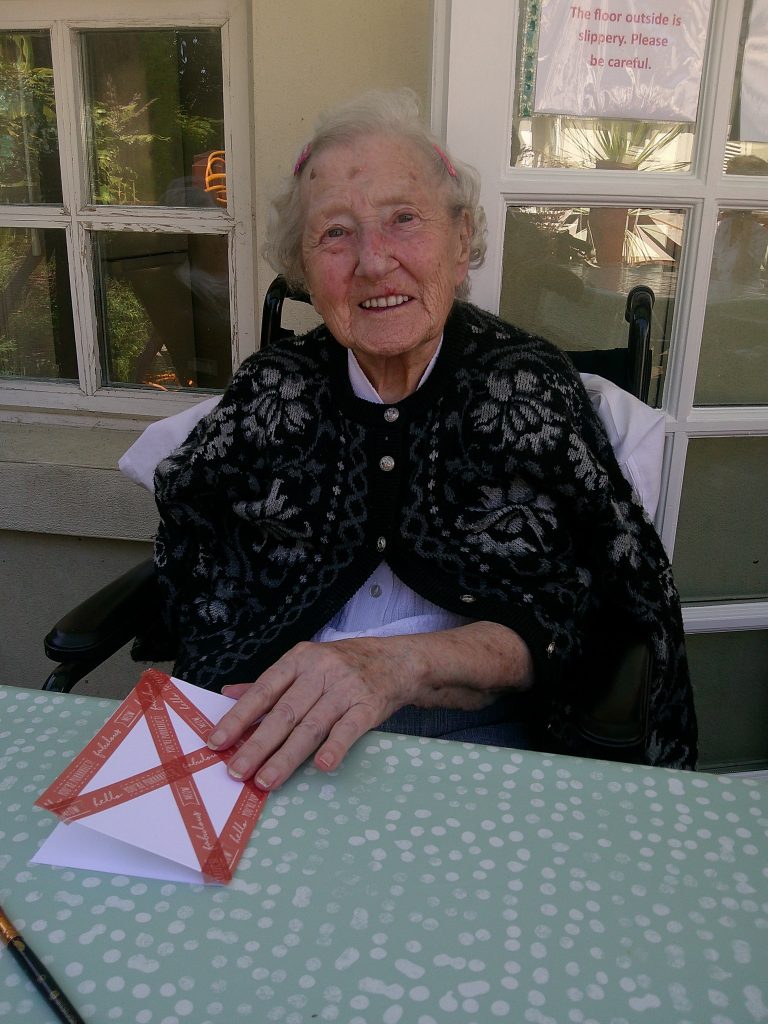 Smiling elderly woman sitting in a wheelchair at a table, holding a folded paper craft outside a building.
