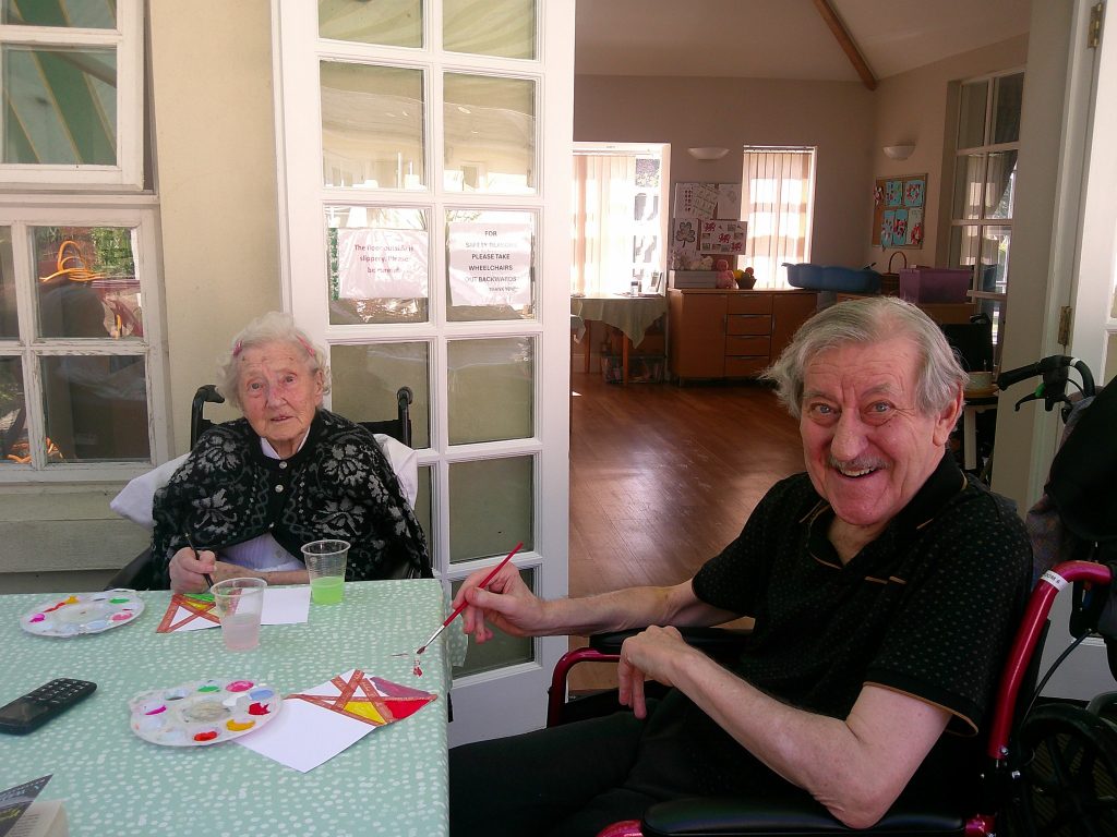 Two elderly residents sitting at a table, smiling and painting colorful paper crafts together in a care home.