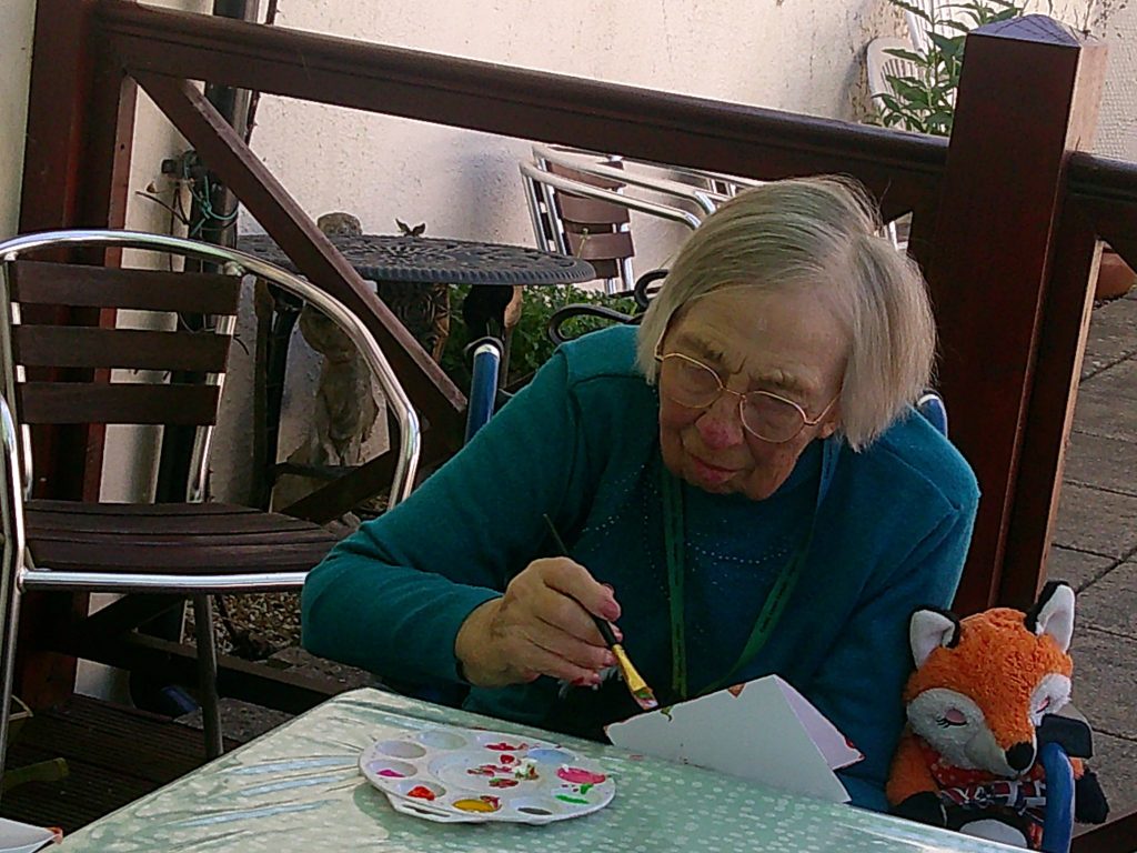 Elderly woman wearing glasses painting a paper craft at a table outdoors, with art supplies beside her.