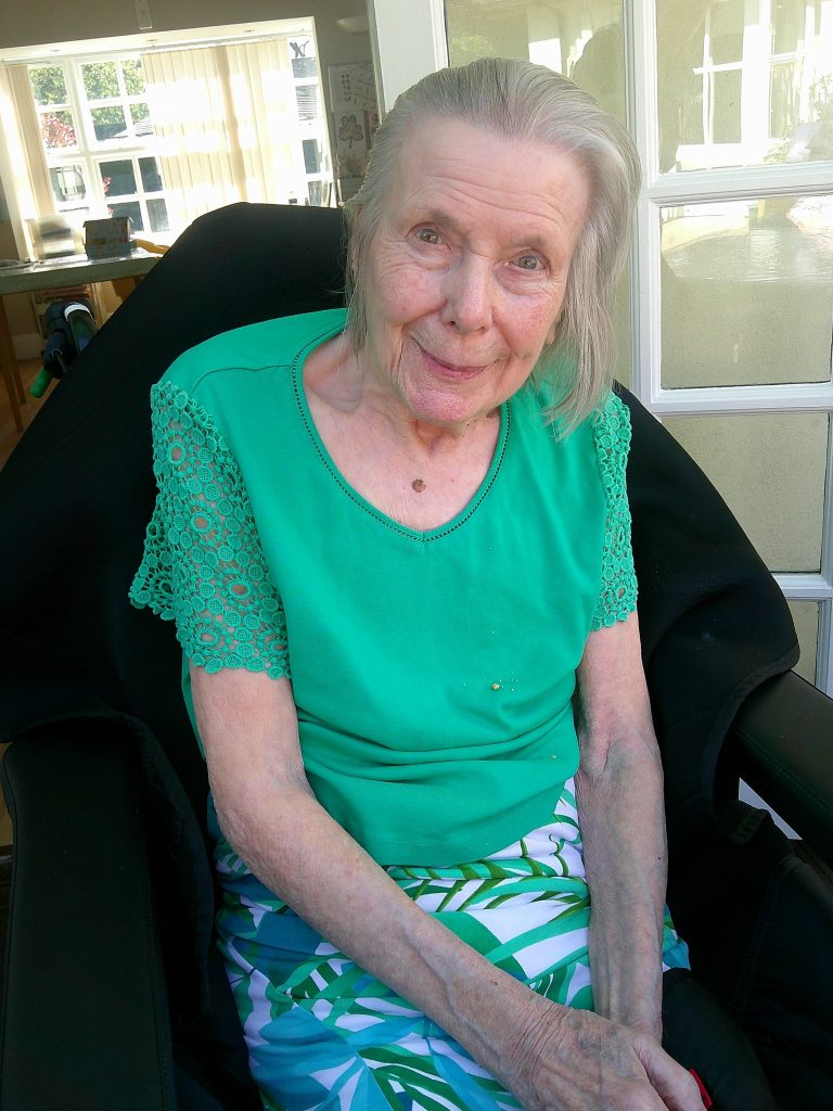 Elderly woman in a green top sitting in a chair indoors, smiling gently.