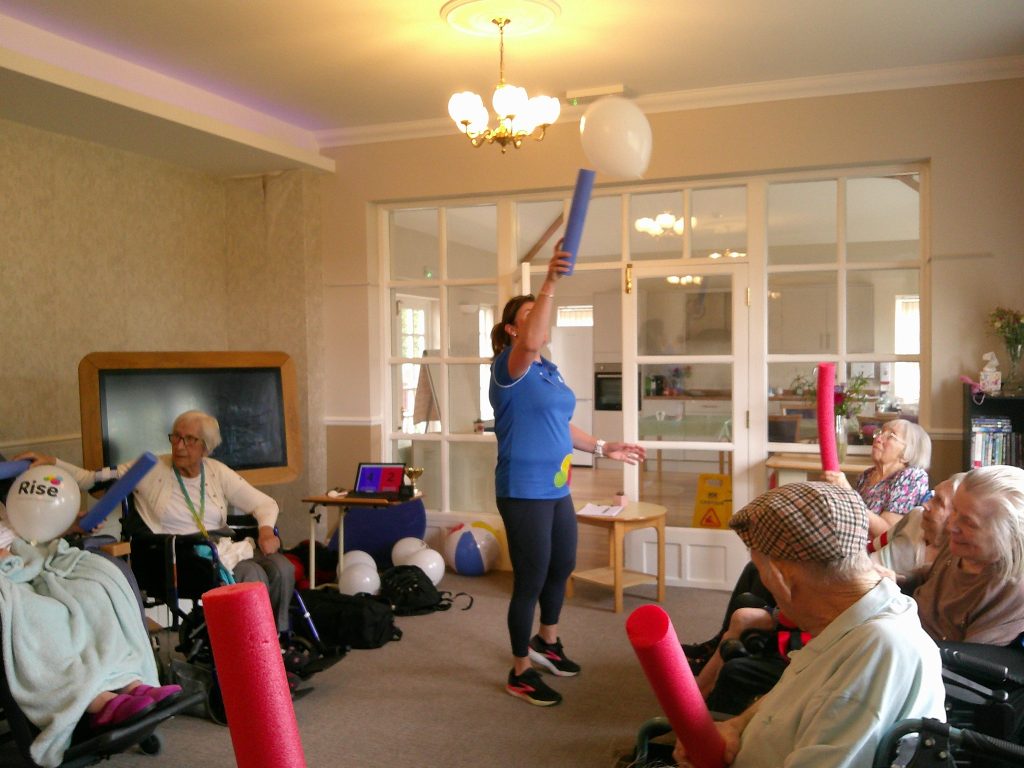 Elderly residents participate in a group activity with a caregiver, using foam sticks and balloons in a care home.