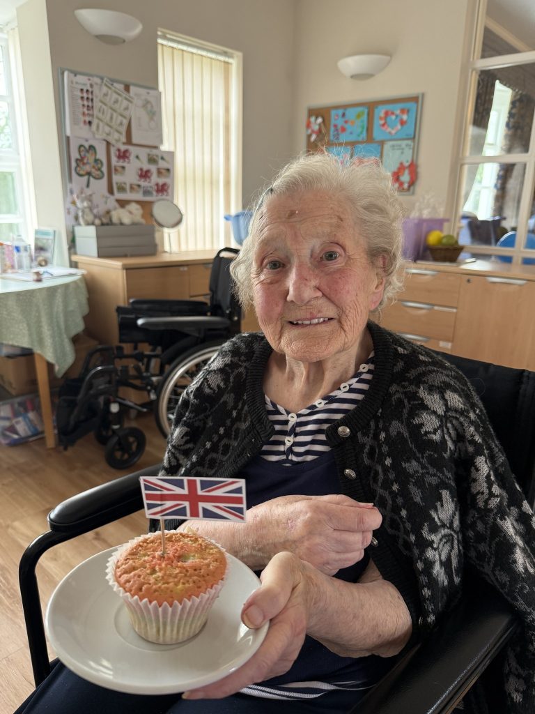 Elderly woman in a wheelchair smiles while holding a cupcake with a small Union Jack flag.