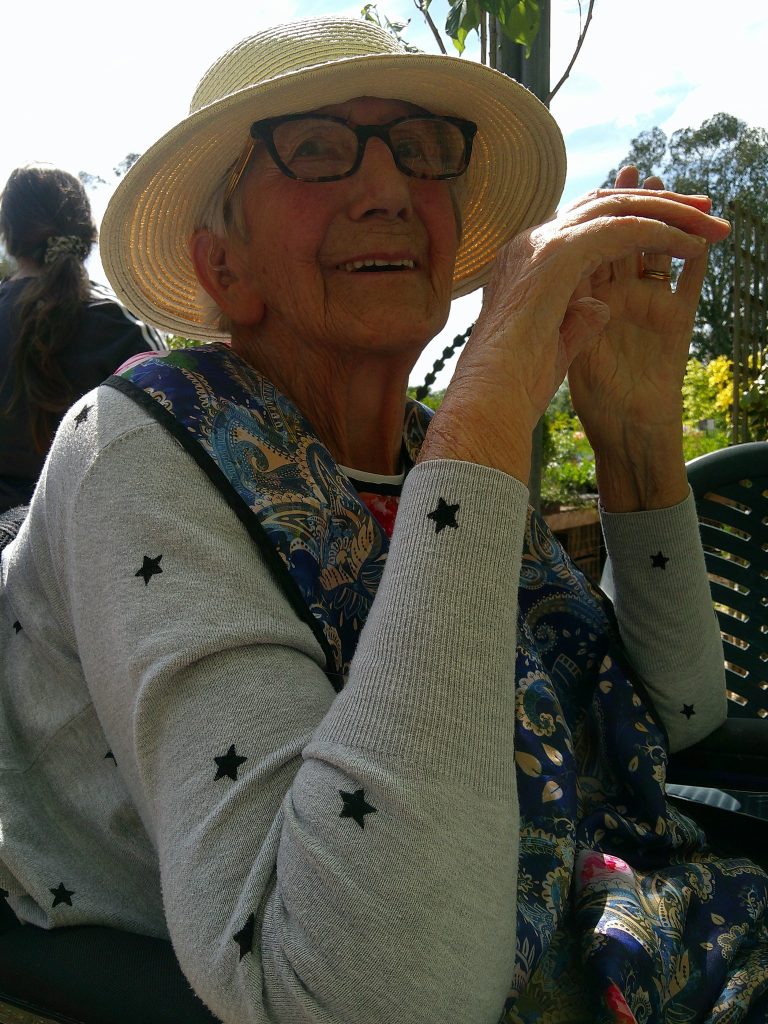 An elderly woman wearing a sunhat and glasses smiles while sitting outdoors in a garden setting.