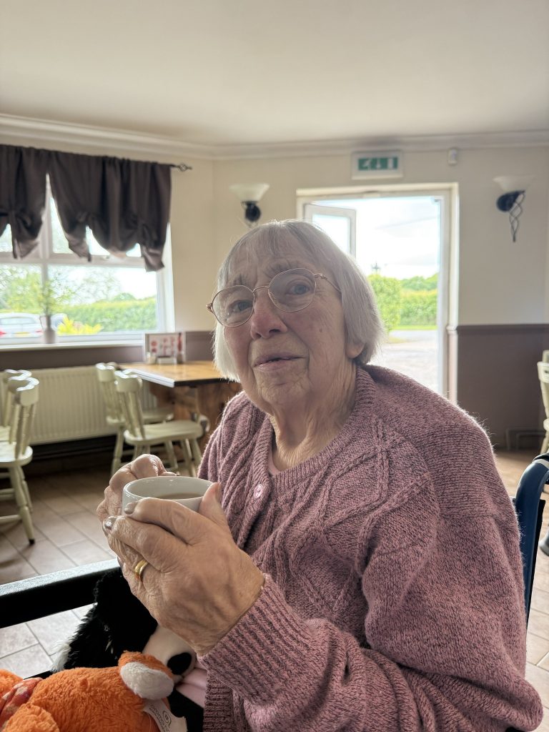 Elderly woman with glasses holds a cup of tea, sitting in a bright café.