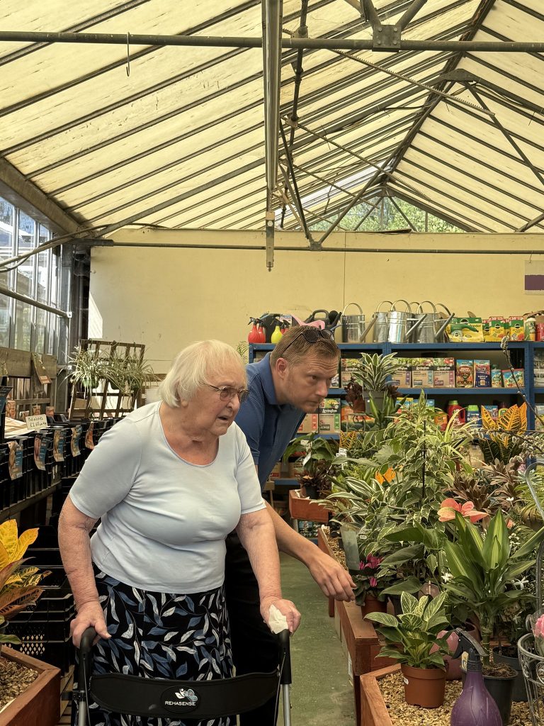 An elderly woman with a walker and a caregiver look at plants inside a garden center greenhouse. 
