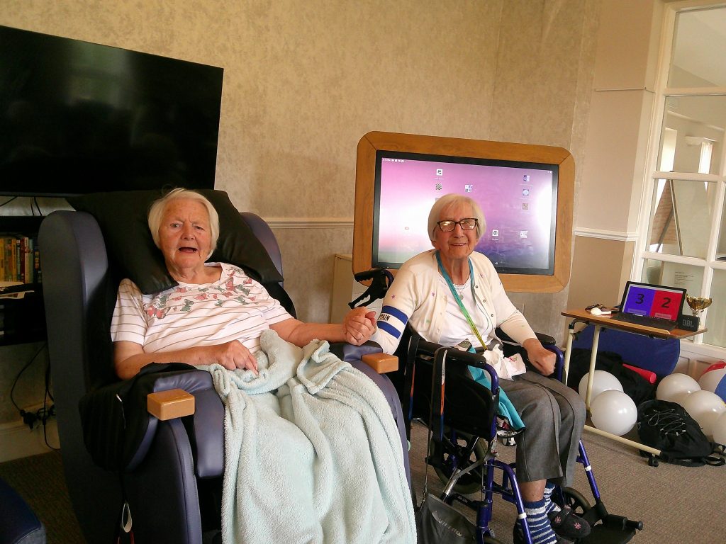 Two elderly women sit together holding hands, one in a wheelchair, in a cozy indoor setting.