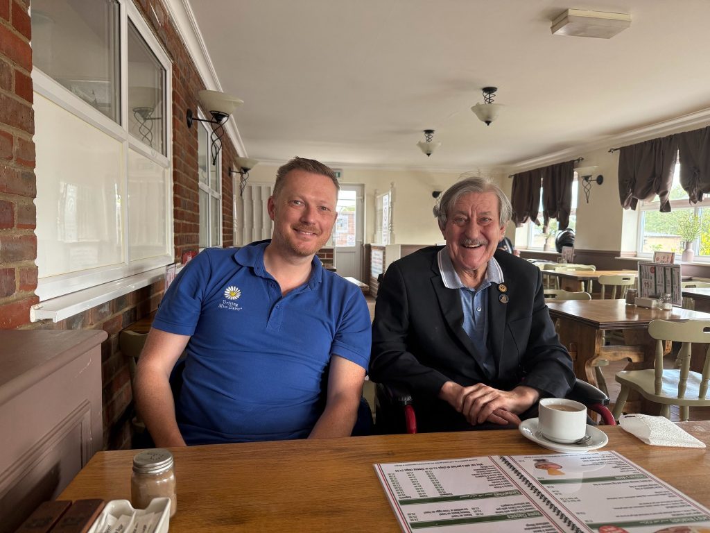 Two men sit at a café table smiling, with a menu and cups of coffee in front of them.