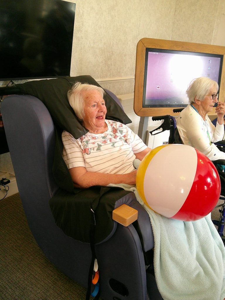 An elderly woman sits in a recliner smiling while holding a colorful beach ball during a recreational activity.