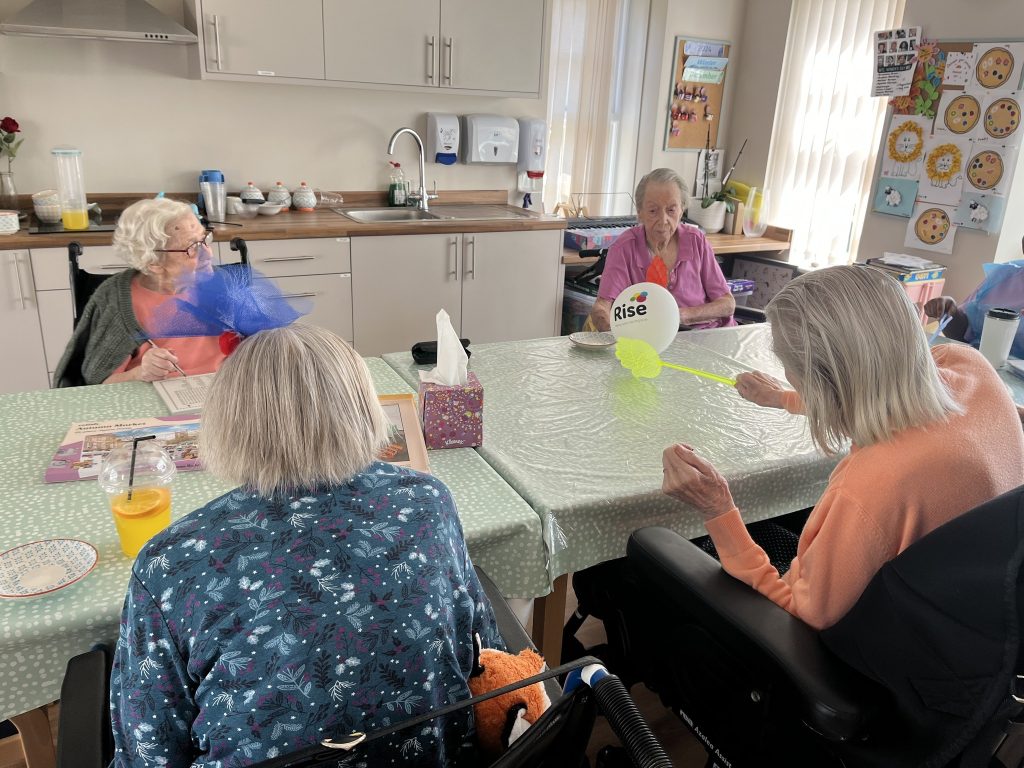 Elderly residents sit around a table playing a balloon game in a care home activity room.