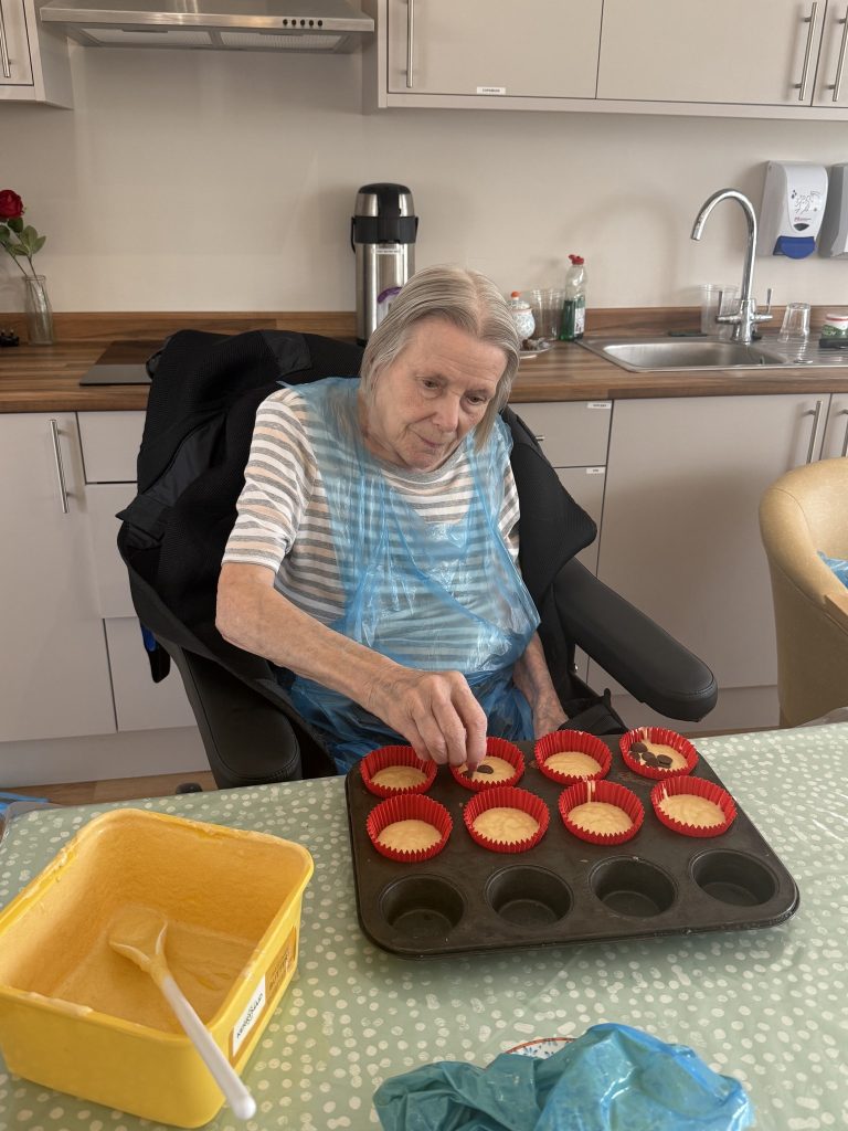 Elderly woman decorating cupcake batter in a care home kitchen.