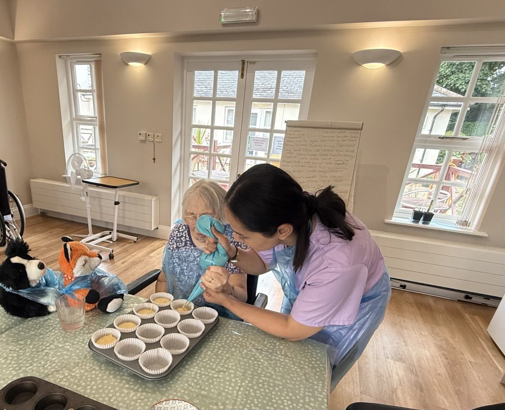 Caregiver helping an elderly woman pipe cupcake batter into paper cases at a table.