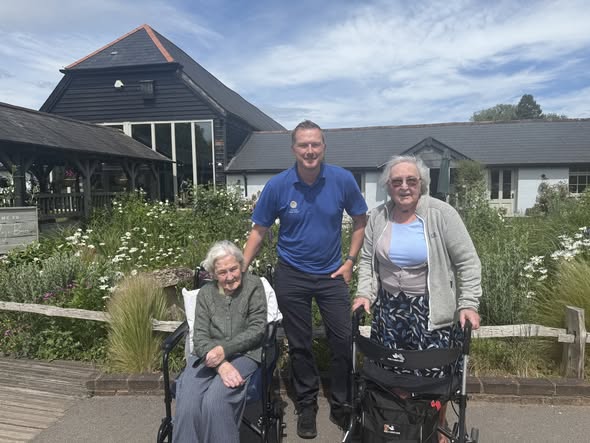 A man stands with two elderly women, one seated in a wheelchair and one using a walker, in front of a care home garden.