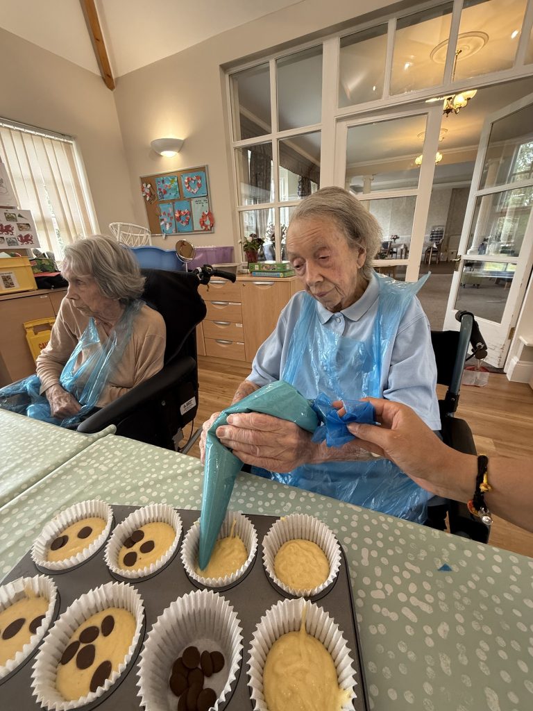 Two elderly women in aprons preparing cupcake batter with assistance in a communal room.