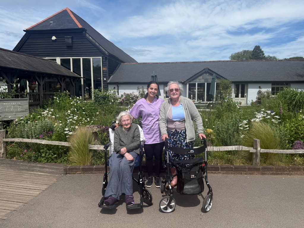 Caregiver stands with two elderly women—one in a wheelchair and one with a walker—outside in a garden area.