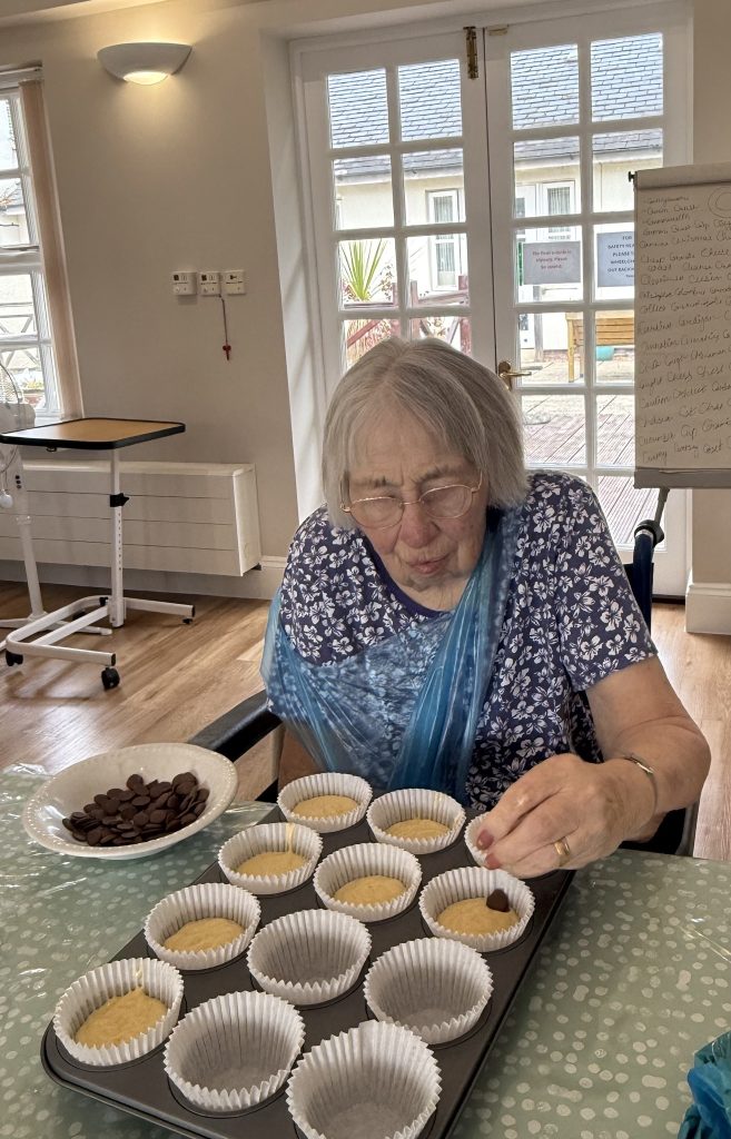 Elderly woman placing chocolate chips into cupcake batter while seated at a table.