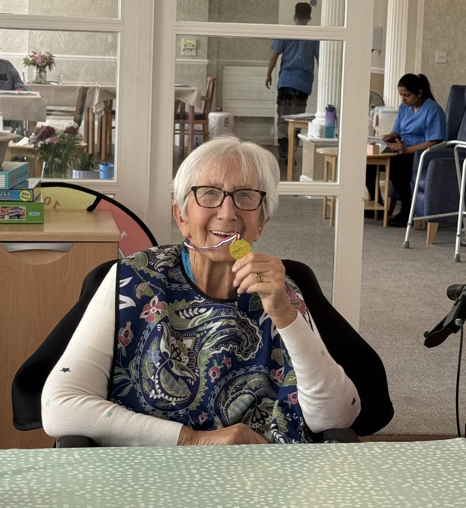 Elderly woman smiling while holding a medal in a care home lounge.