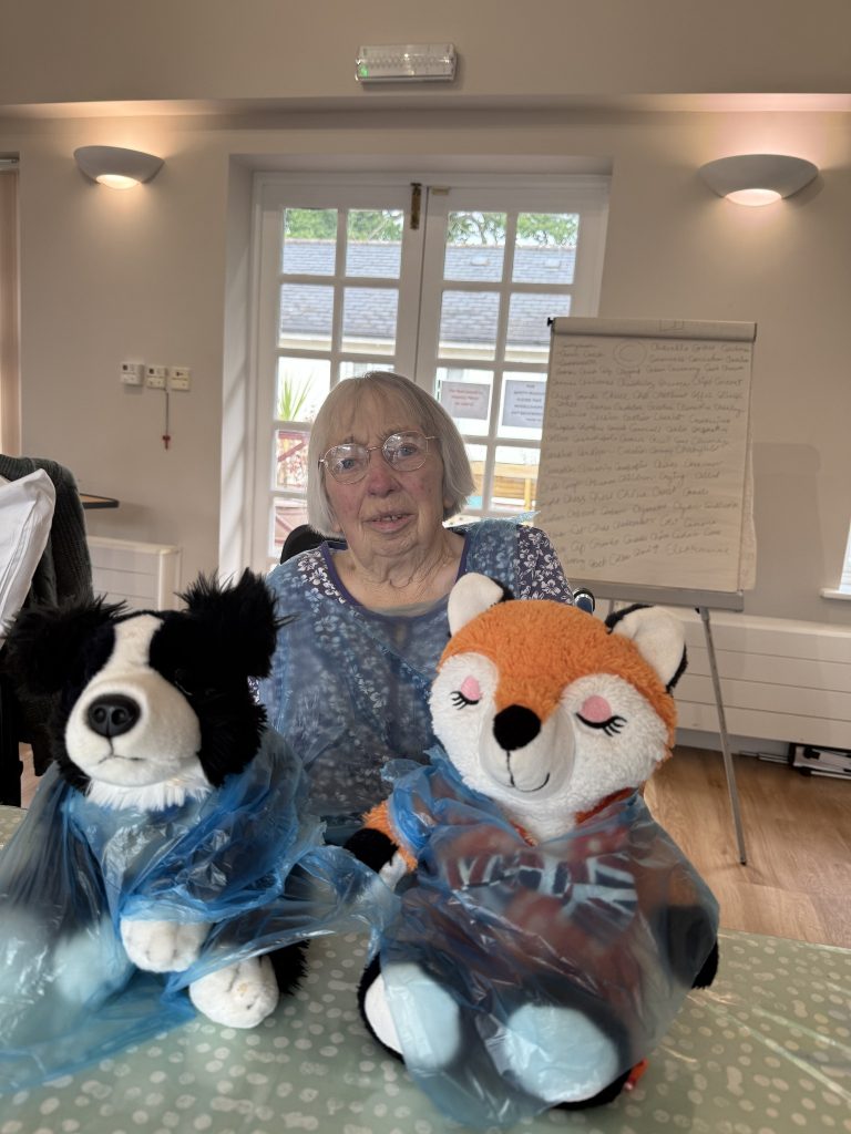 Elderly woman sitting with two plush toys wearing aprons at a table in a care home.