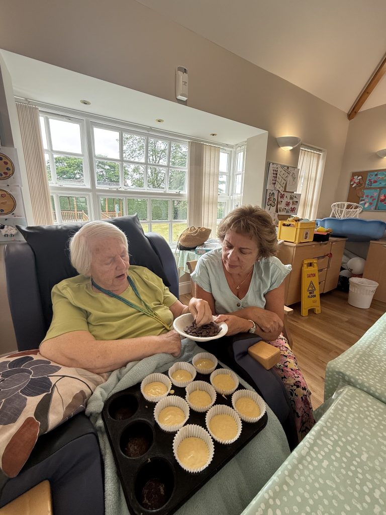 Elderly woman and caregiver adding chocolate chips to cupcake batter in a bright care home room.