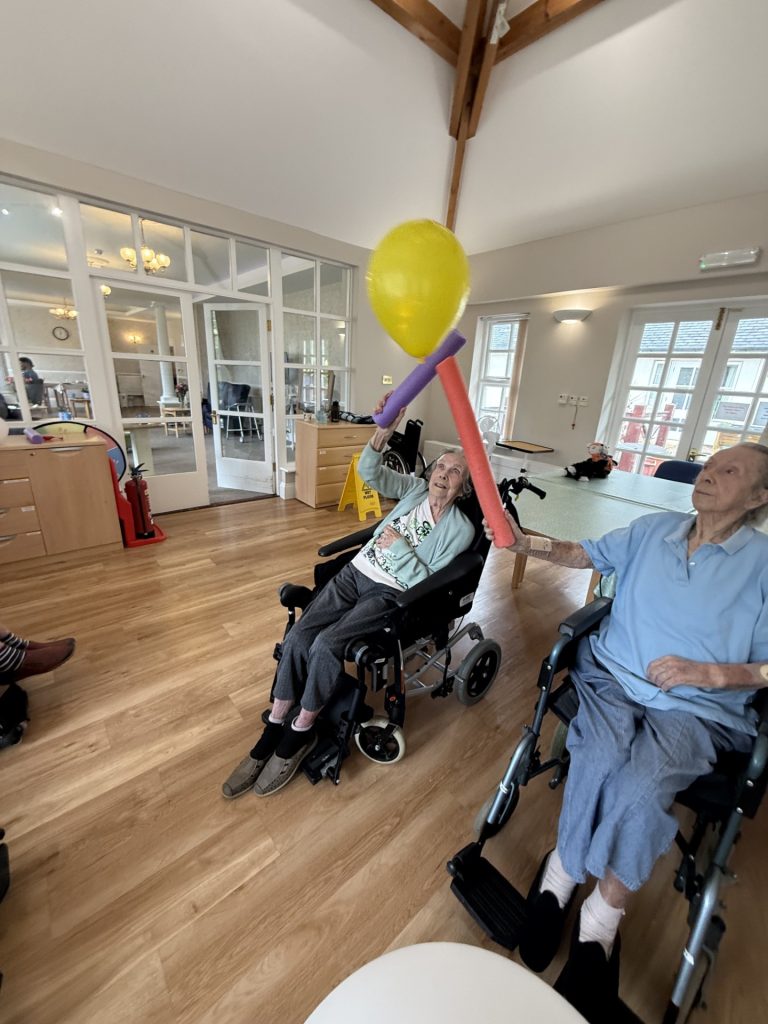 Two elderly people in wheelchairs reach up to tap a yellow balloon during a group activity.