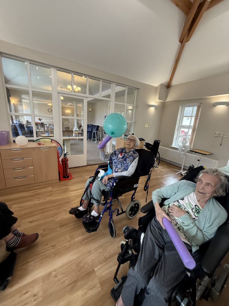 Seniors in wheelchairs smile and play a balloon game with foam sticks in a care home lounge. 