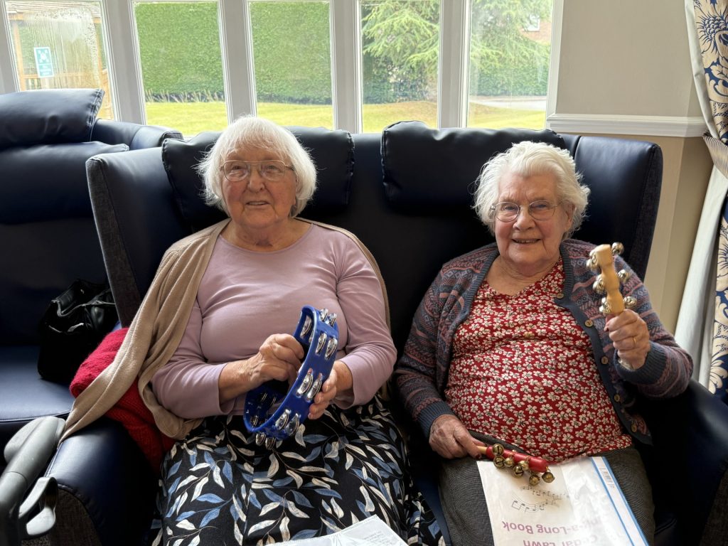 Two elderly women sitting on a sofa, smiling and holding tambourines.
