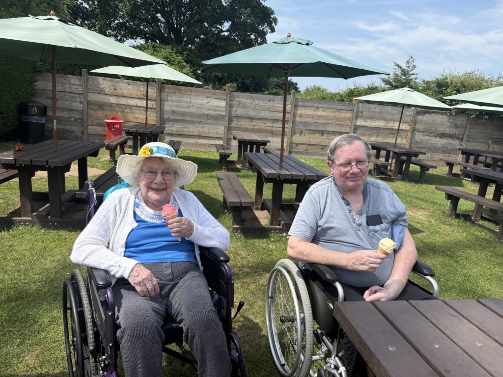 Two elderly people in wheelchairs sitting outdoors, smiling and holding ice creams.