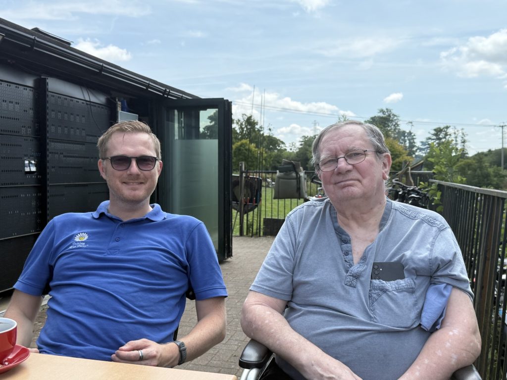 Two men sitting outside at a table, one in a wheelchair, smiling at the camera.
