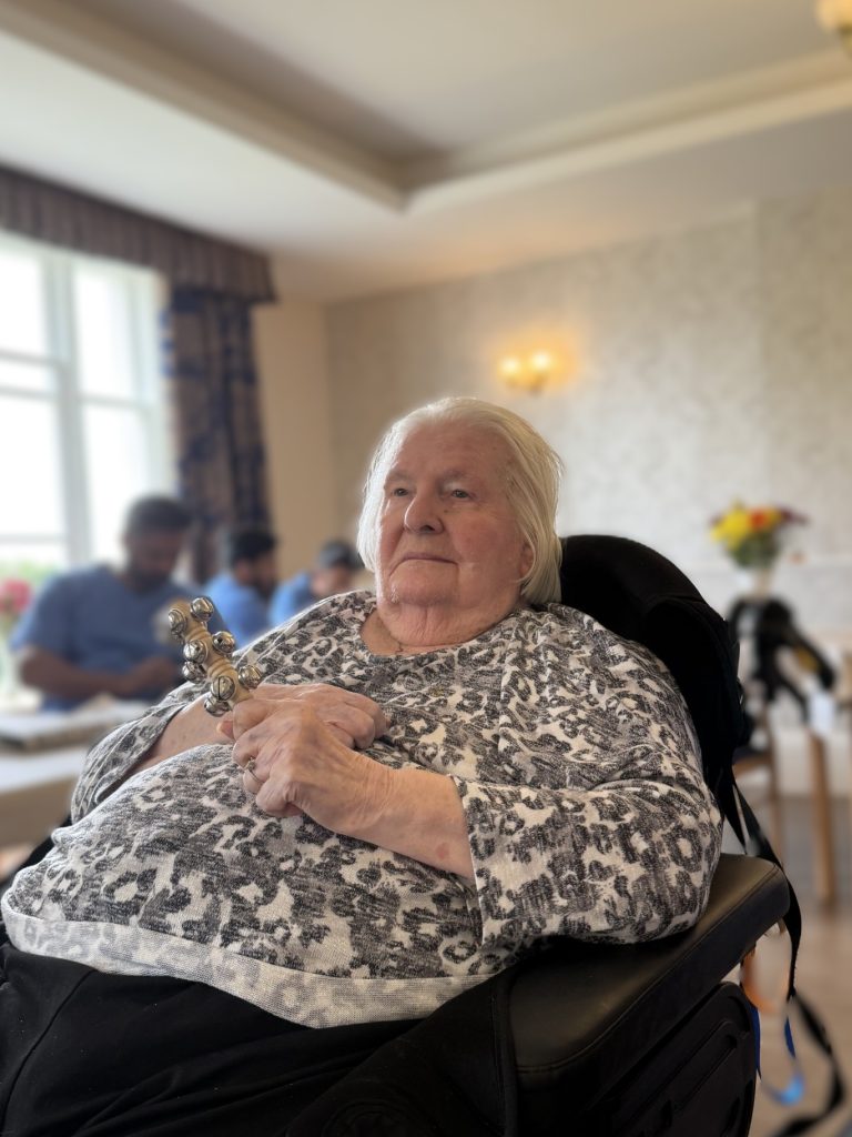 Elderly woman sitting in a chair indoors, holding a small jingle bell instrument.