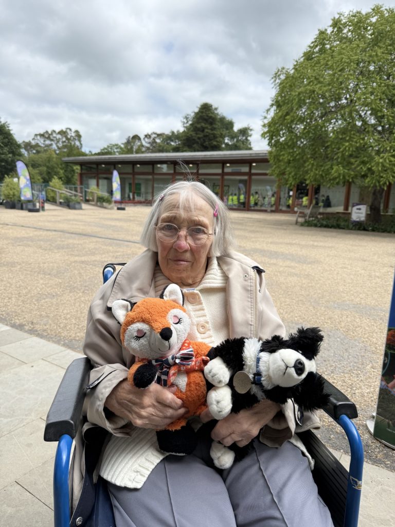 An elderly woman in a wheelchair holding two stuffed animals outside.