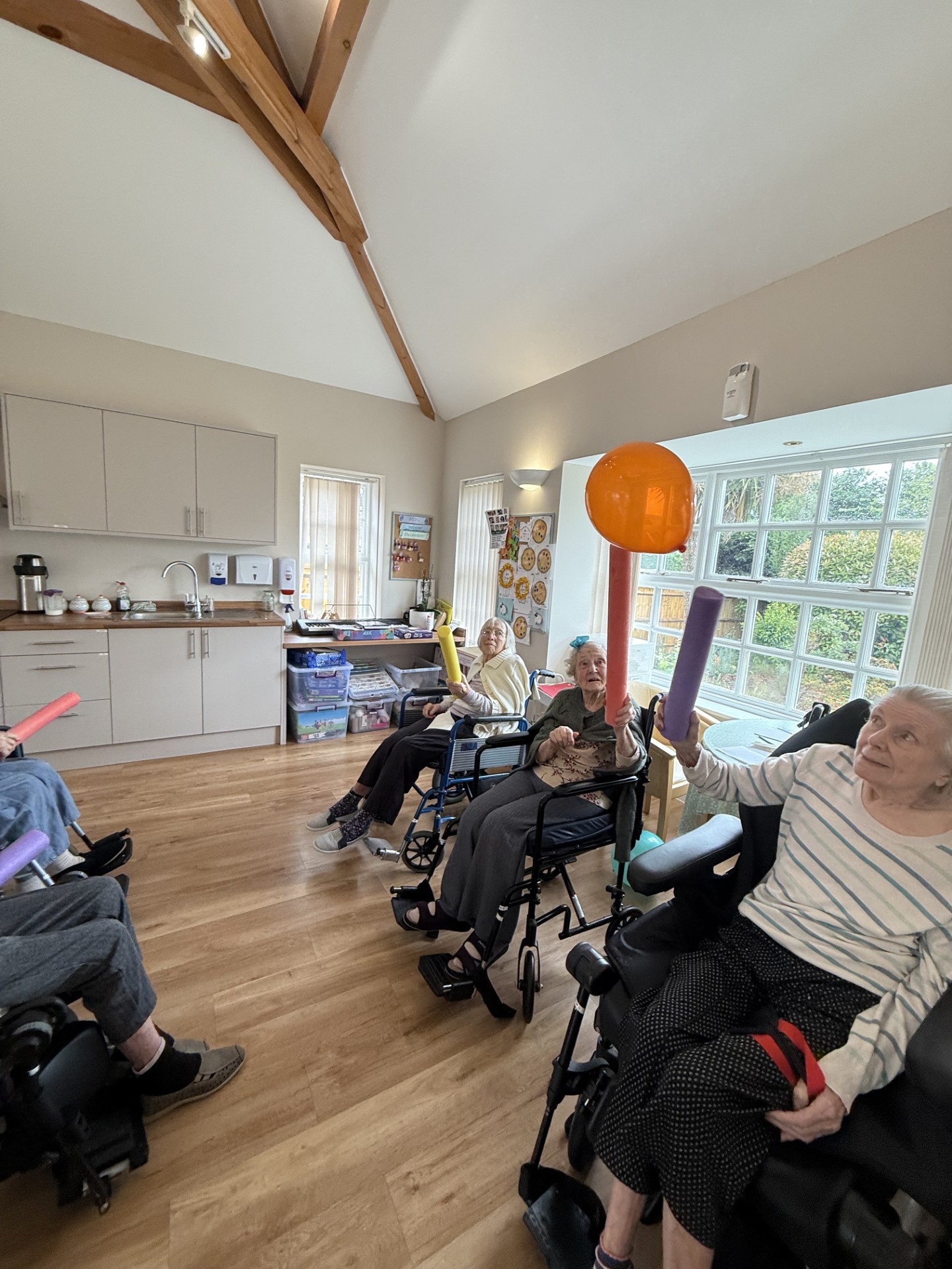 Elderly residents in wheelchairs play with balloons using foam sticks in a bright communal room.