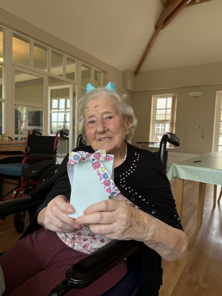 An elderly woman in a wheelchair holding a small decorated gift box indoors.