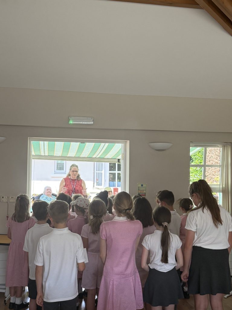 Group of schoolchildren standing indoors, watching a woman speaking at the front.