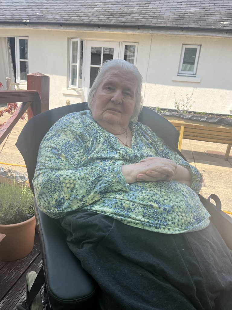 Elderly woman seated in a chair outside, wearing a patterned top, looking calmly at the camera.