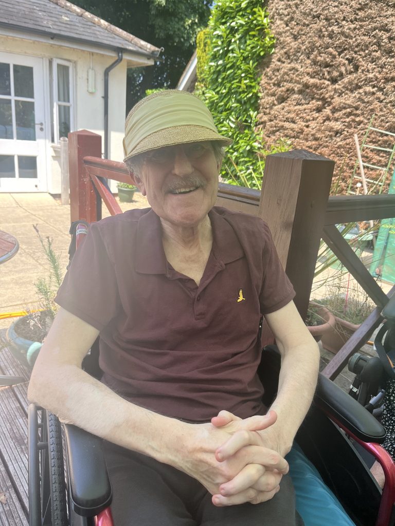 Elderly man in a hat sitting in a wheelchair outdoors, smiling with hands clasped.